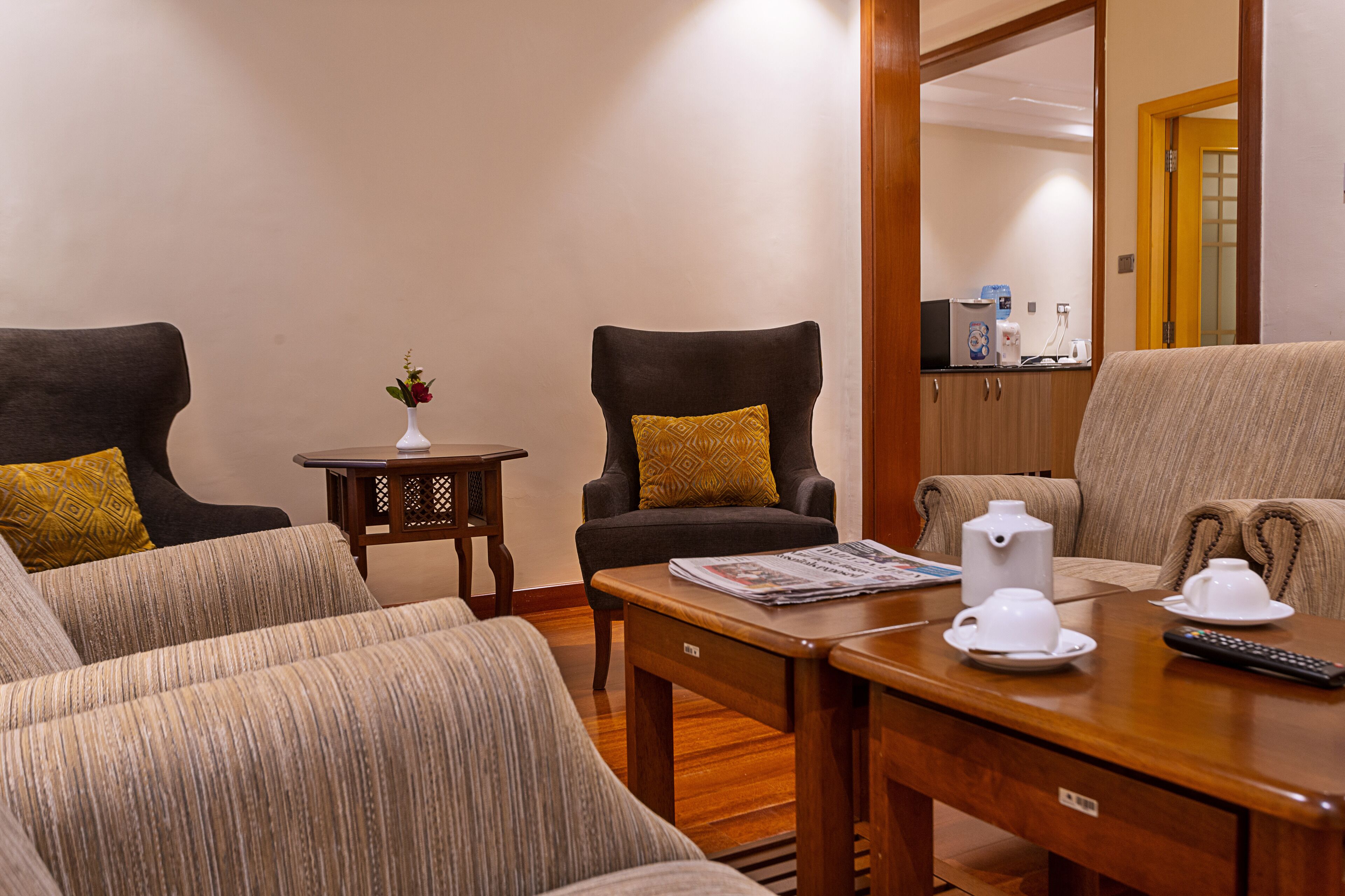 A hotel room with a sofa set, a small table with a vase of flowers, and a kitchen area visible through an open door.