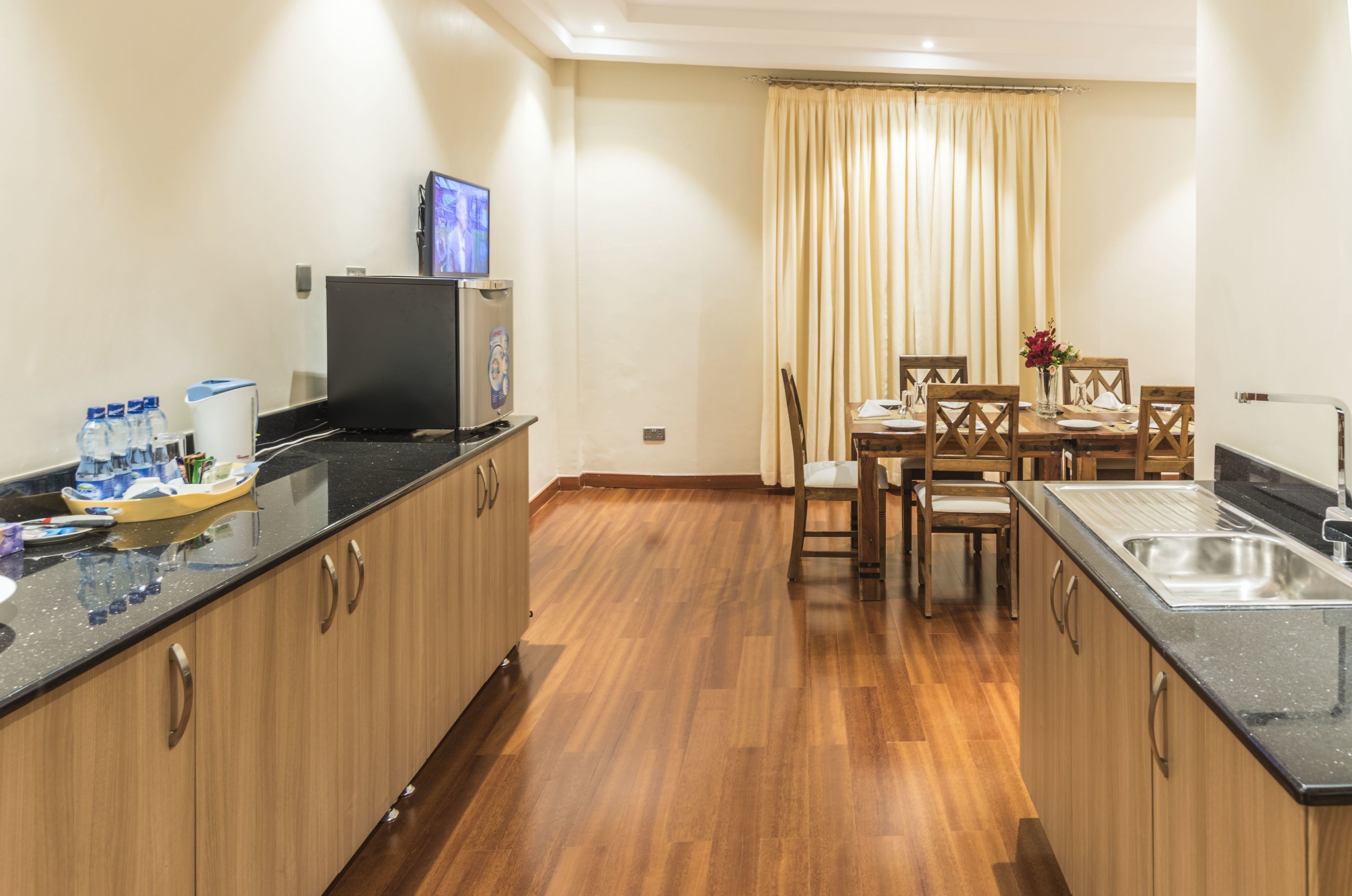 A modern kitchen with wooden cabinets, a black countertop, a dining area with a table and chairs, and a TV mounted on the wall.