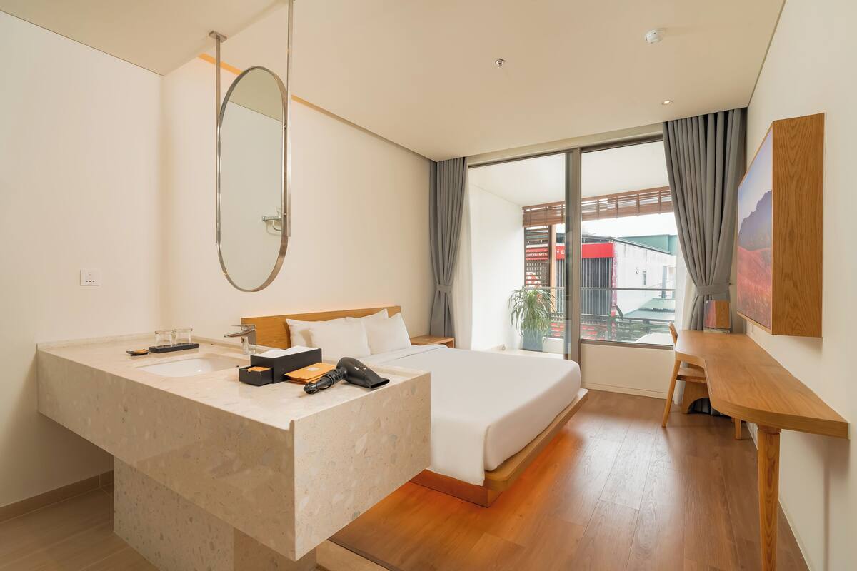 A modern hotel room with a large bed, a built-in vanity with a mirror, a wooden desk, and a large window offering a view of the outside.