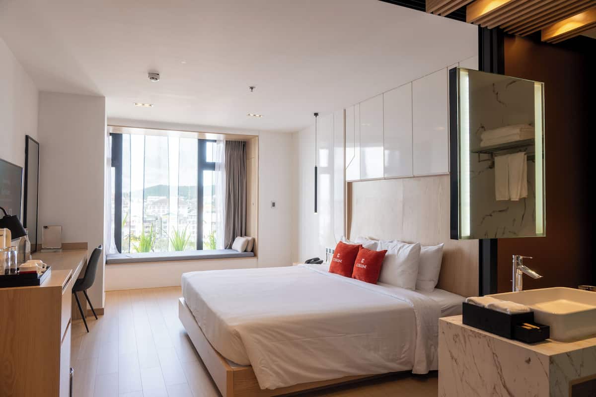 A modern hotel room with a large bed, a desk, a bathroom with a mirror, and a window with a view of the city.