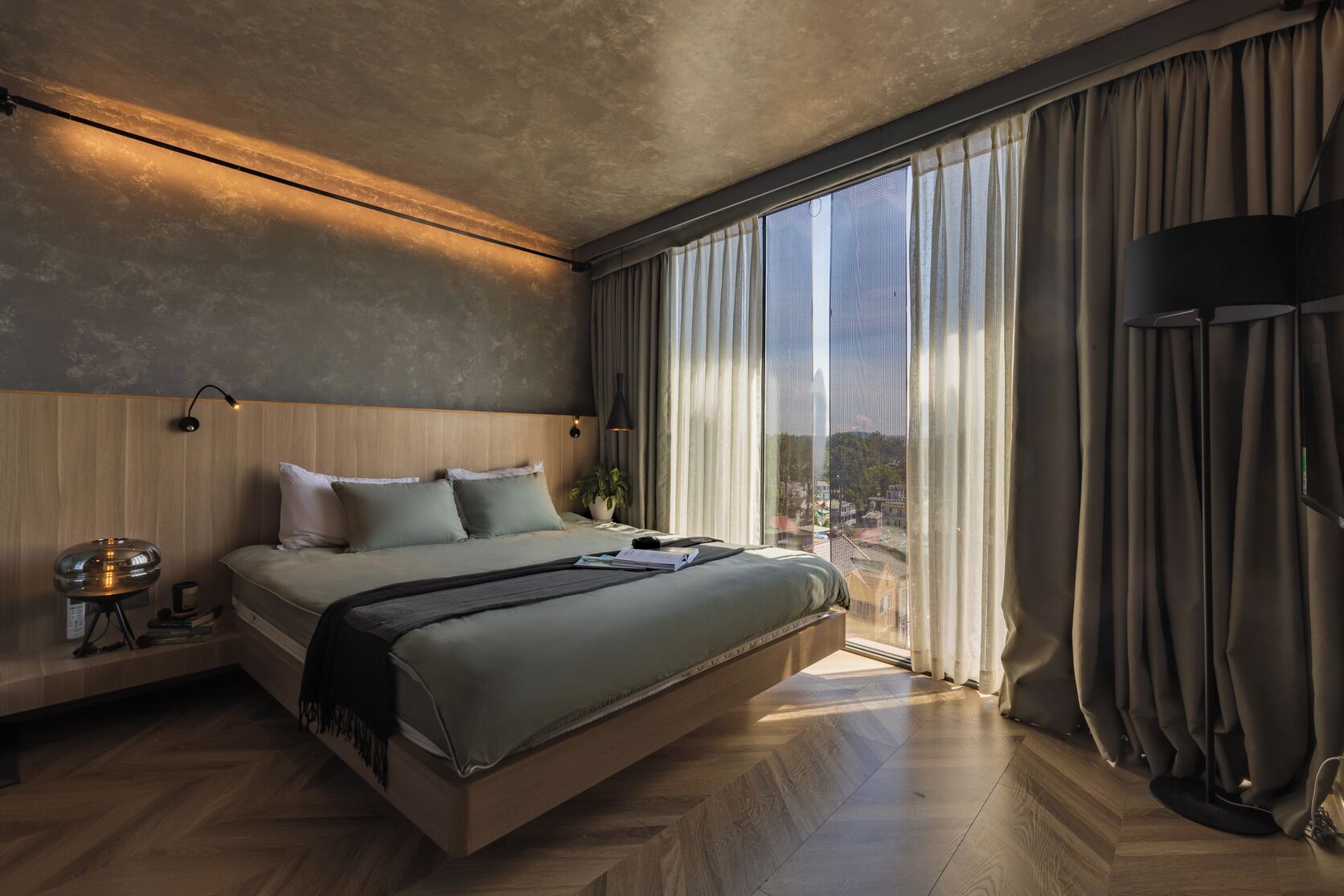Un dormitorio moderno con una cama grande, una mesita de noche, una lámpara y vistas de la ciudad a través de amplios ventanales.