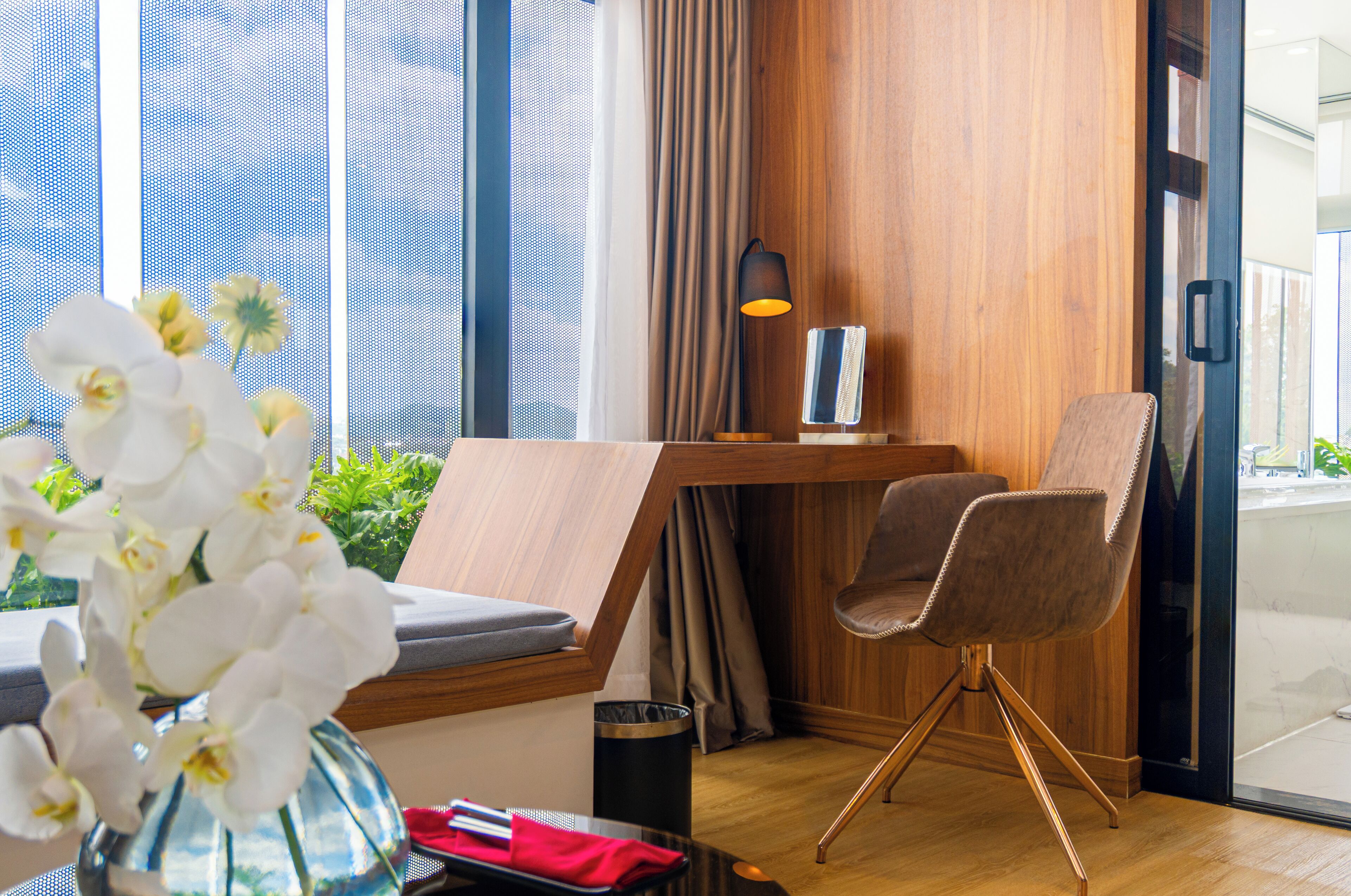 Une chambre d’hôtel moderne avec un bureau en bois, un fauteuil confortable, un vase rempli d’orchidées blanches et une vue sur l’extérieur grâce à de grandes fenêtres.