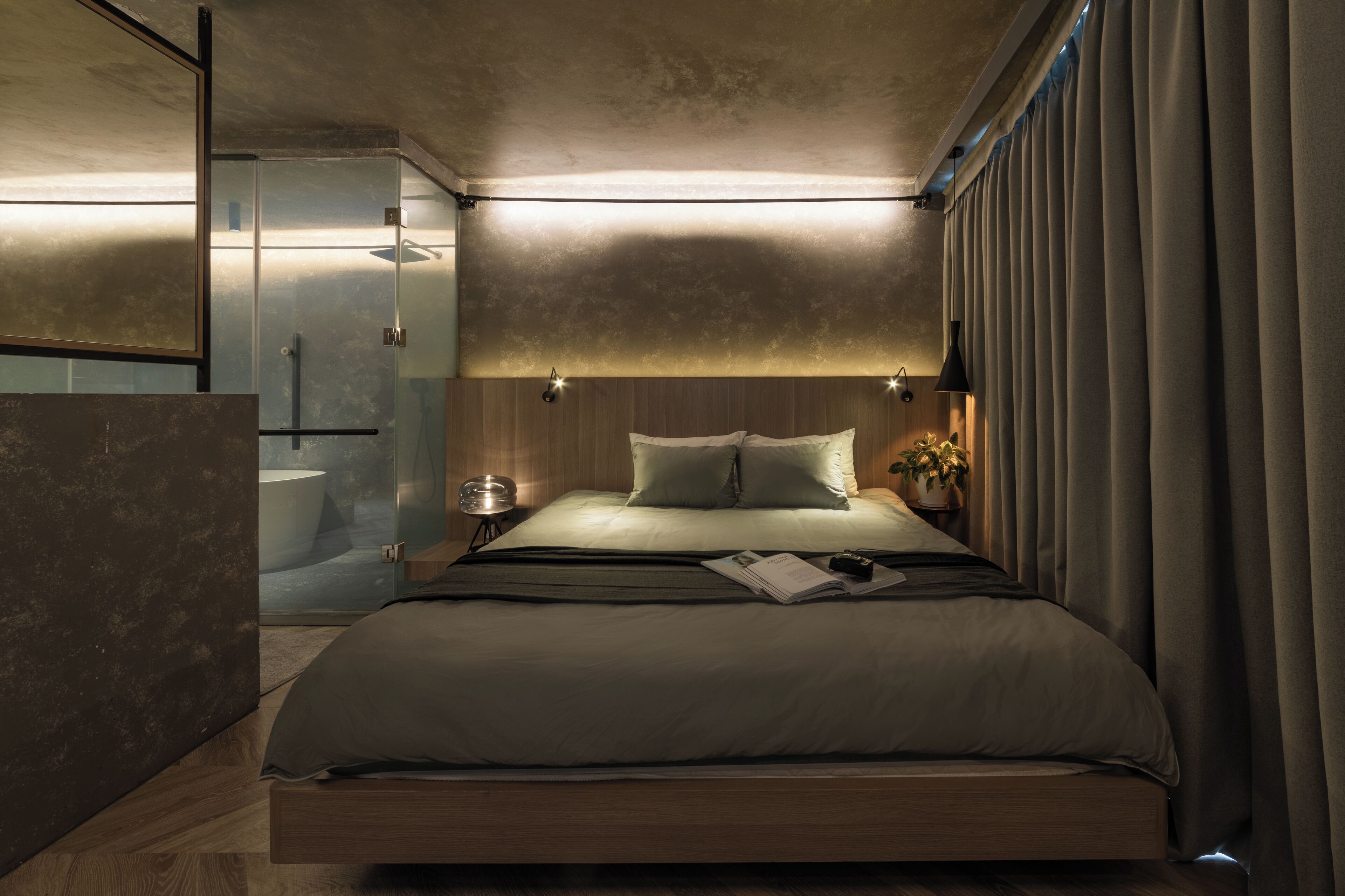 A modern bedroom with a large bed, bedside lamps, a nightstand, and a potted plant.