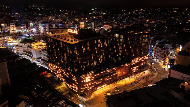 A multi-story hotel with illuminated signage, a parking area, and surrounding urban buildings. - Colline Dalat (Xuan Huong - Da Lat)