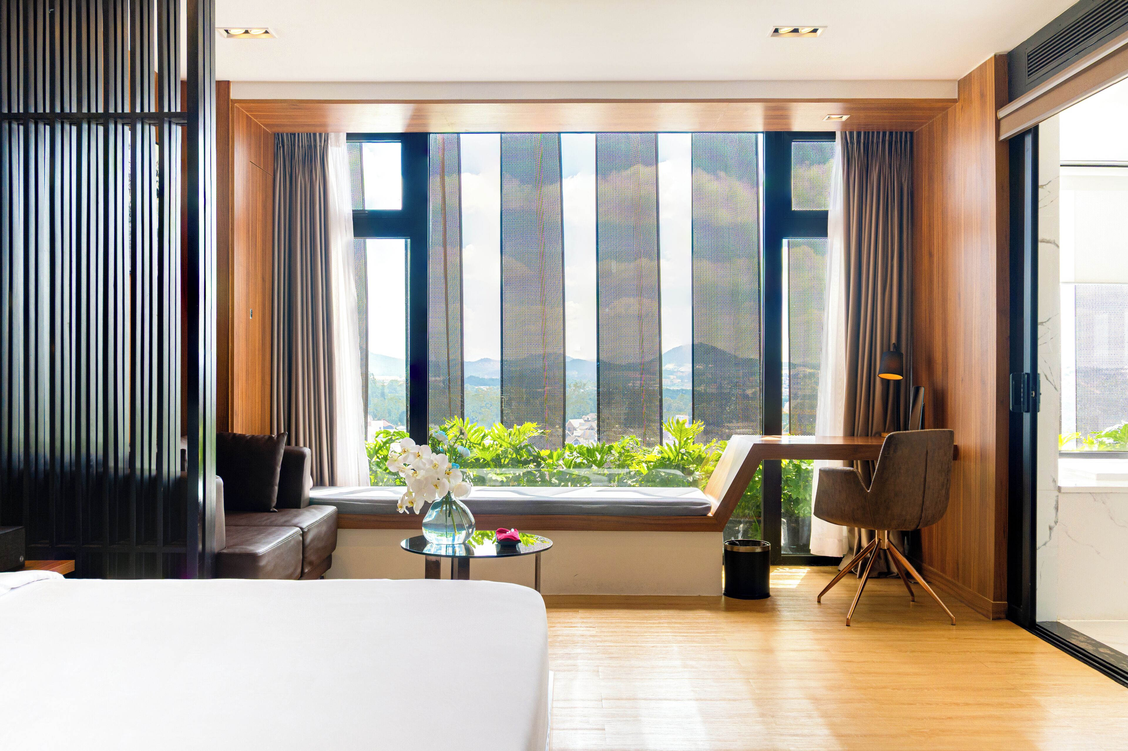 A hotel room with a large window, a bed, a sofa, a small table with a vase of flowers, and a chair.