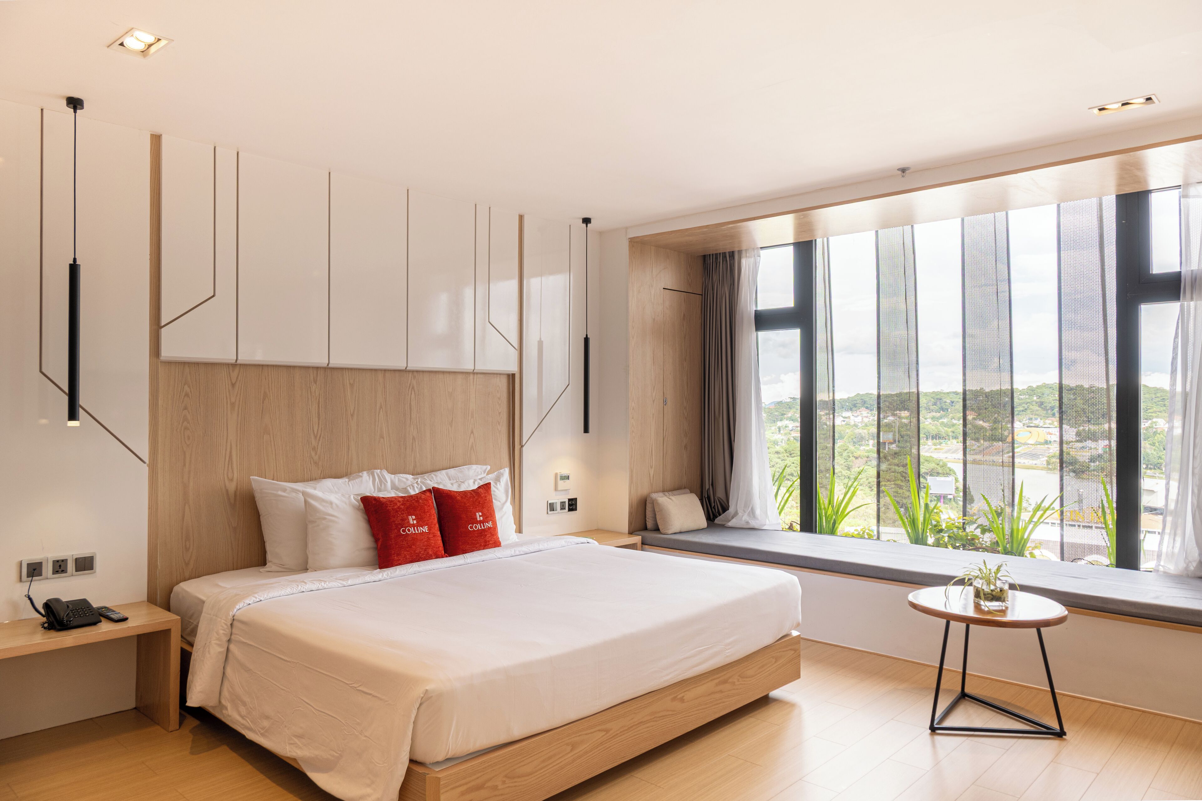A modern bedroom with a large bed, a bedside table, a small round table, and a window with a view of greenery.