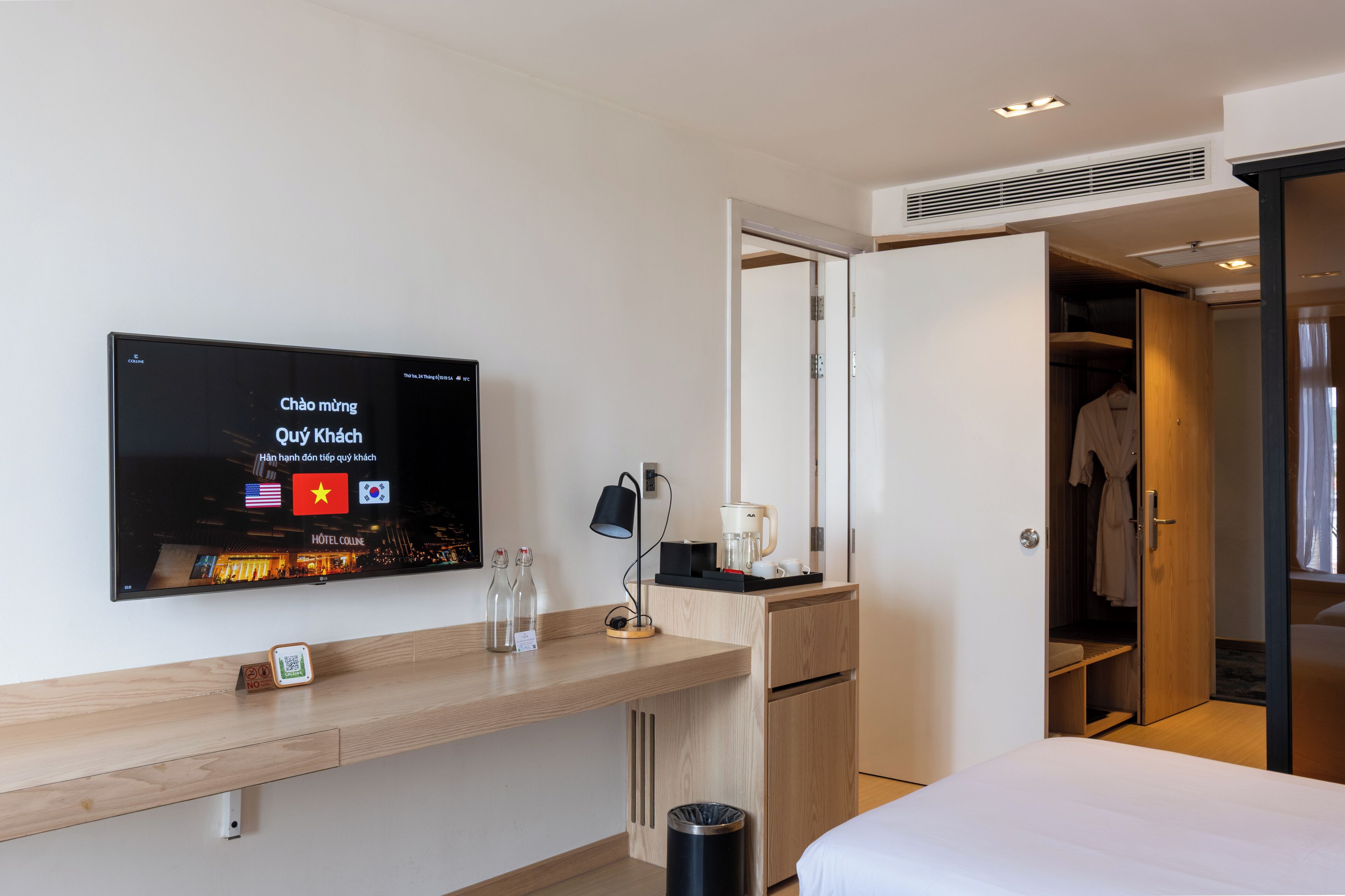 A hotel room with a bed, a desk with a television displaying hotel information, a coffee maker, and a wardrobe.