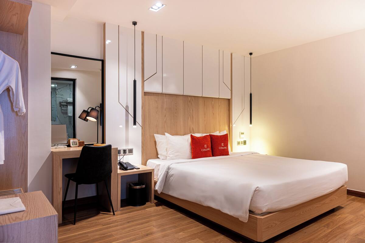 A modern hotel room with a wooden bed frame, a desk with a chair, a small bedside table, and a mirror.