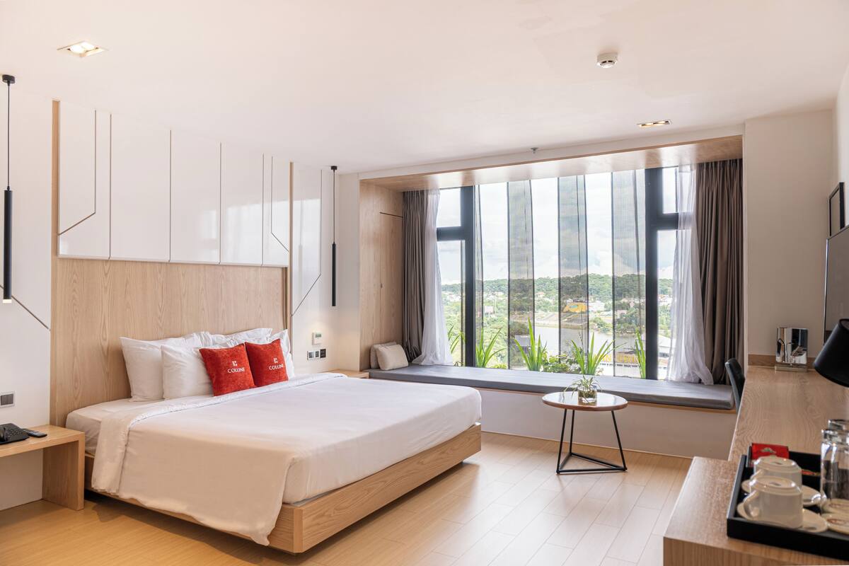 A modern hotel room with a large bed, a small table with a plant, a desk, and a view of the city through large windows.