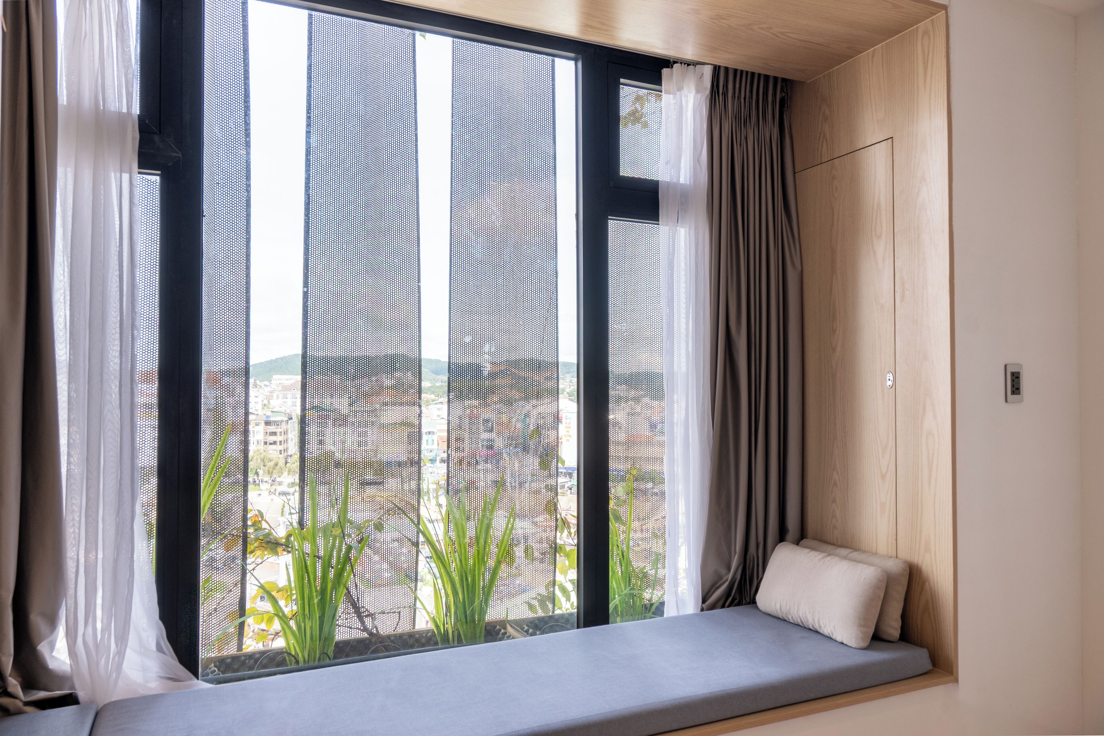 Quarto com uma janela ampla, cama com travesseiro e vista para edifícios e plantas lá fora.
