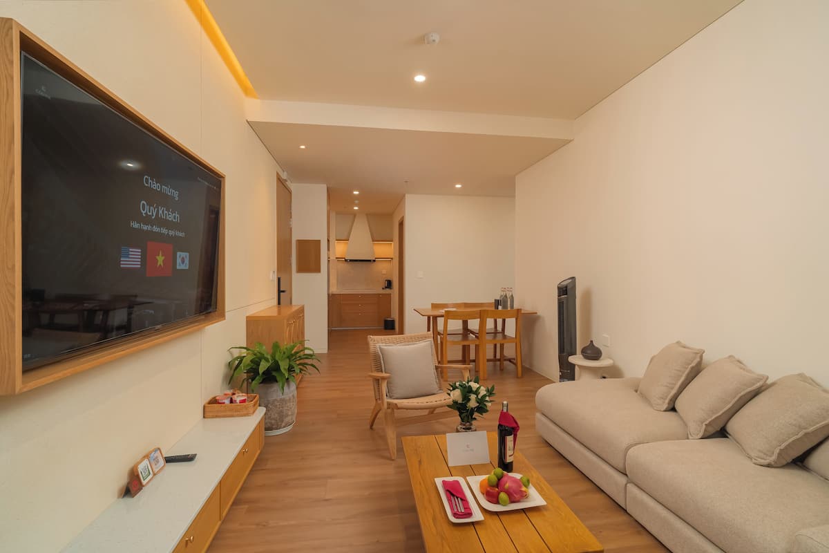 A modern living room with a sofa, a wooden coffee table with fruits and a bottle, a TV mounted on the wall, and a dining area with a table and chairs.