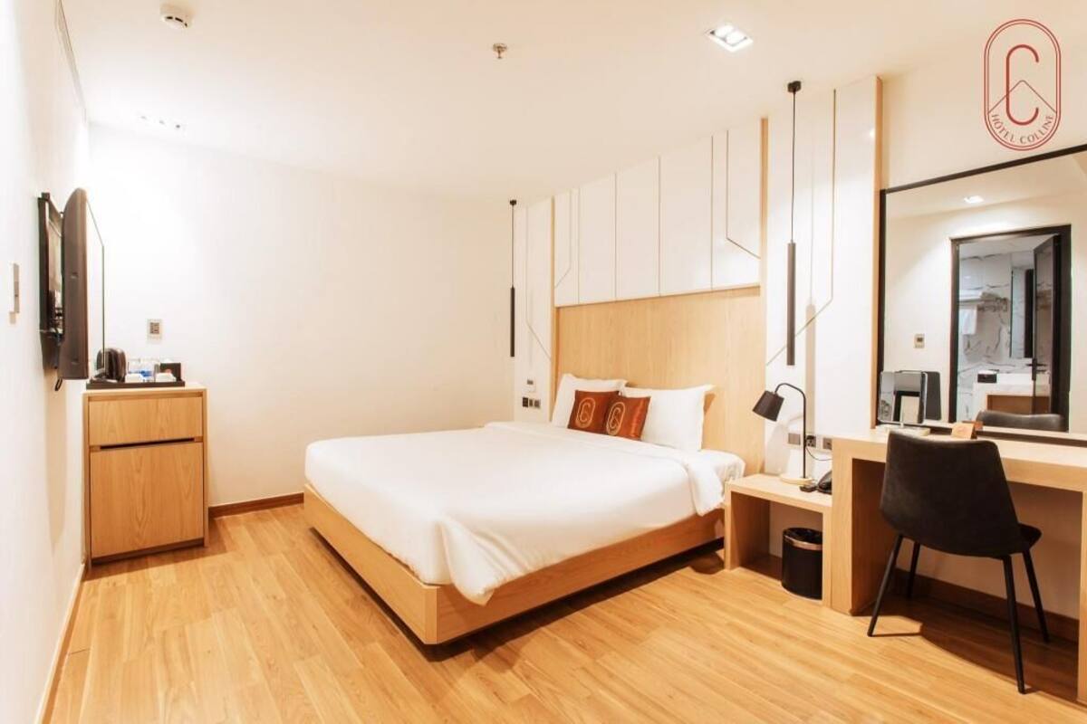 A modern hotel room with a wooden bed, a desk, a chair, a small bedside table, a mirror, and a wardrobe.