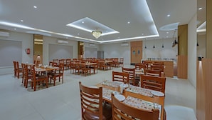 Un vaste espace de salle à manger avec plusieurs tables et chaises, un lustre, des spots encastrés au plafond et, au fond, un coin cuisine.