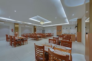 A spacious dining area with multiple tables and chairs, a chandelier, recessed ceiling lights, and a kitchen area in the background.