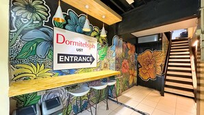 A hotel reception area with a colorful floral mural, a sign indicating the entrance, and a staircase leading to the upper floor. - Dormitelsph UST (Manila)