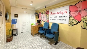 A reception area with a sign reading "DormitelspH" and a floral mural. There are blue chairs, a reception desk, and a water cooler. - Dormitelsph UST (Manila)