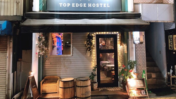 Et hostel med et skilt, der viser "TOP EDGE HOSTEL", samt en lille indgang med en dør. Udenfor står der træfade og en fletstol.