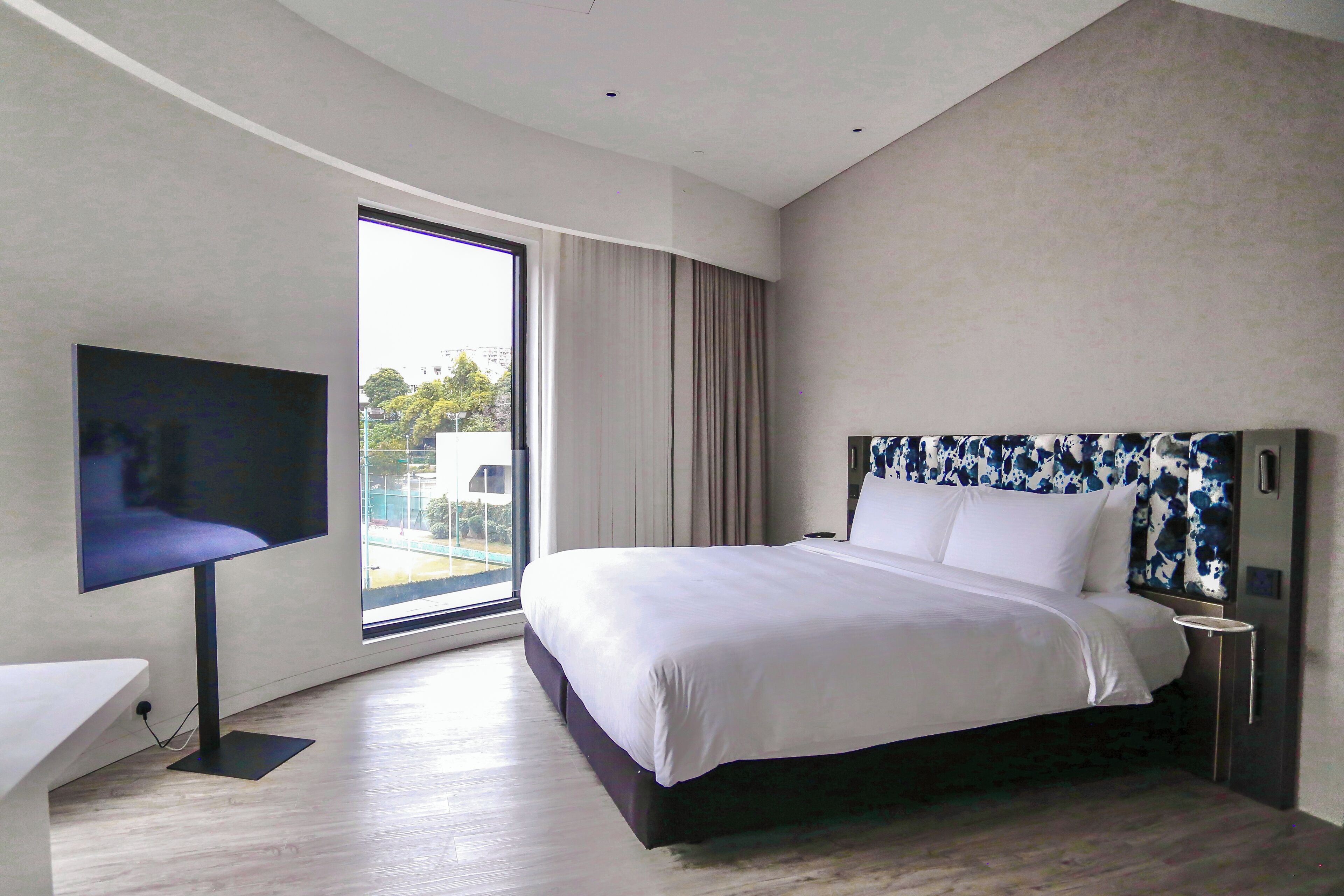 A modern hotel room with a large bed, a flat-screen TV, and a view of the outdoors through a large window.