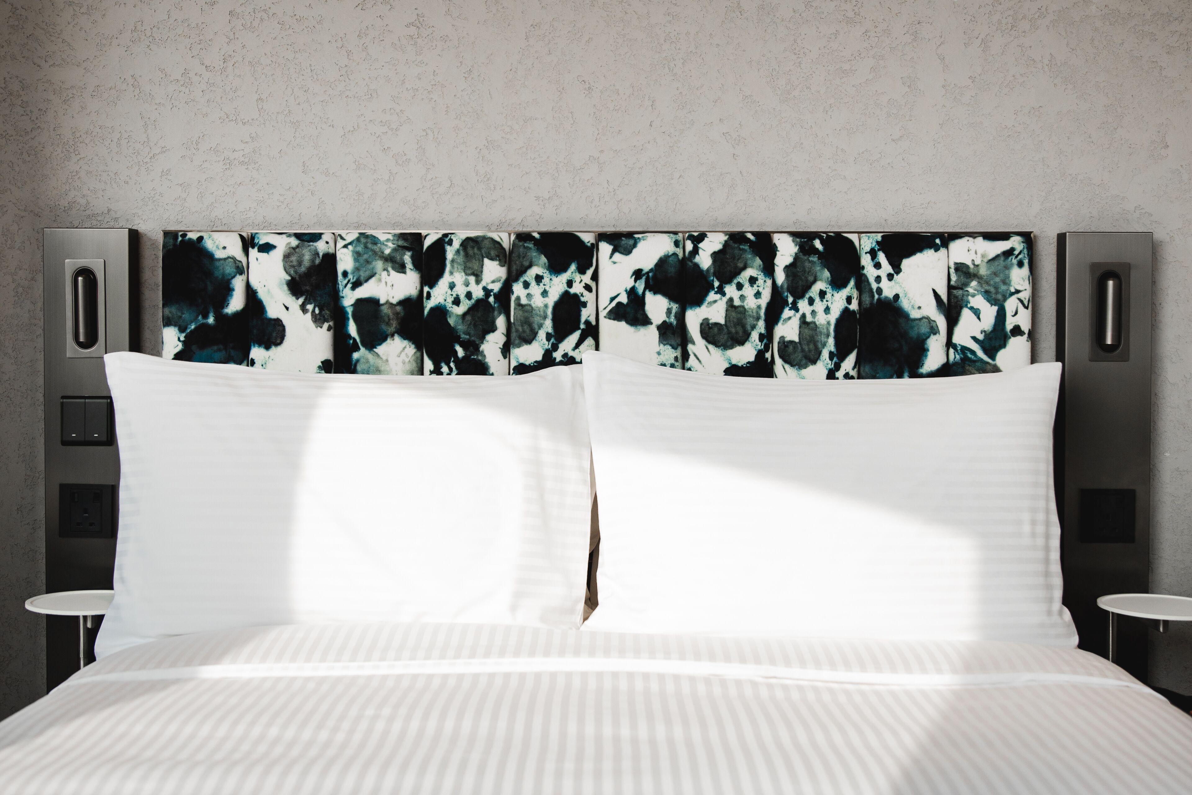 Quarto de hotel moderno com uma cama grande, cabeceira com arte abstrata e duas mesas de cabeceira.