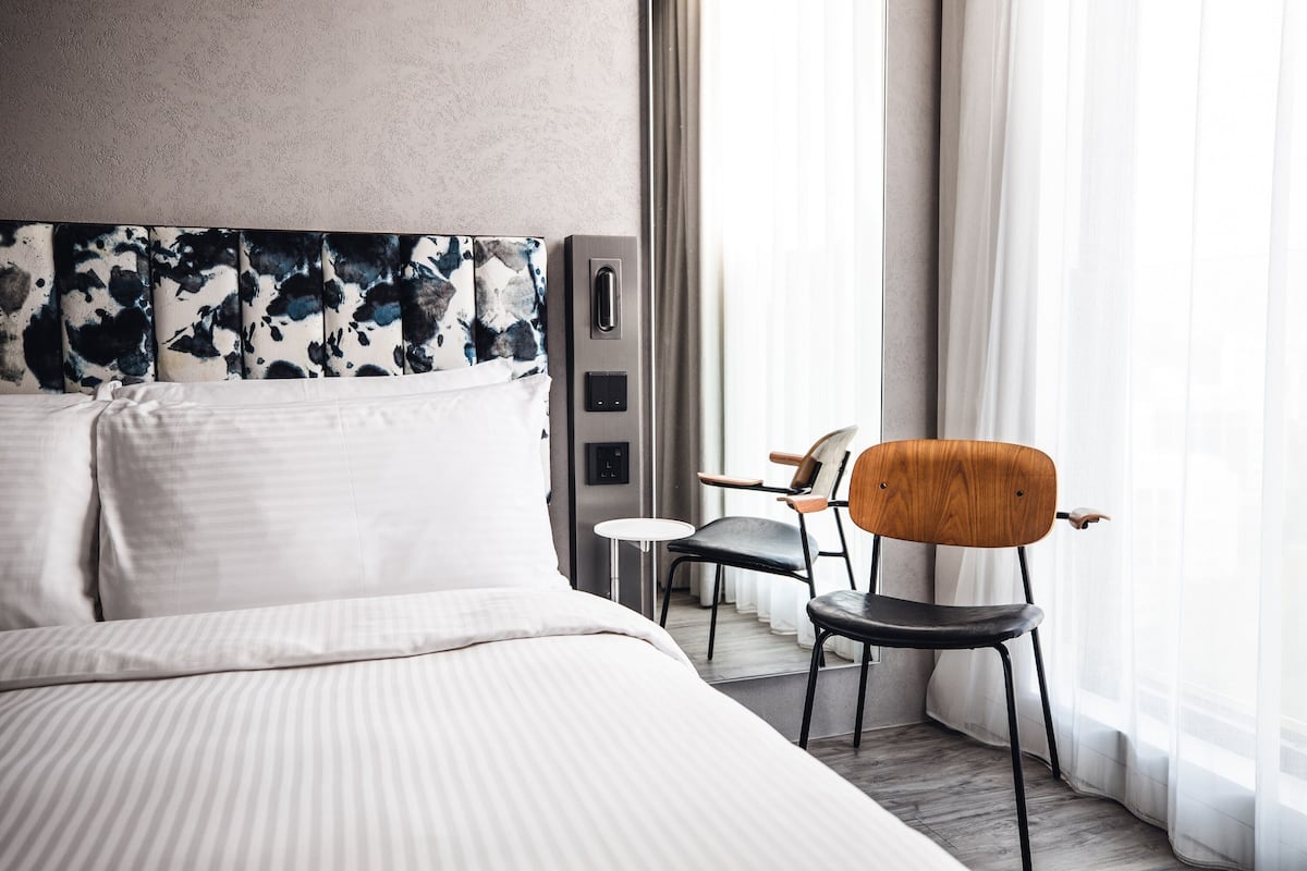 Habitación de hotel con una cama grande, un cabecero moderno, una mesita pequeña, una silla de madera y vistas al exterior a través de cortinas translúcidas.