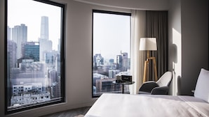 A hotel room with a large window offering a city view, a bed, a chair, a lamp, and a small table with a radio.