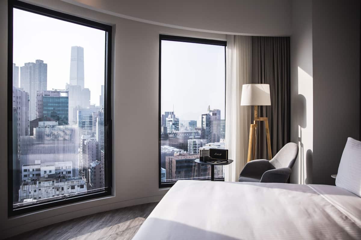 Habitación de hotel con un ventanal grande que ofrece vistas a la ciudad, una cama, una silla, una lámpara y una mesita con un radio.