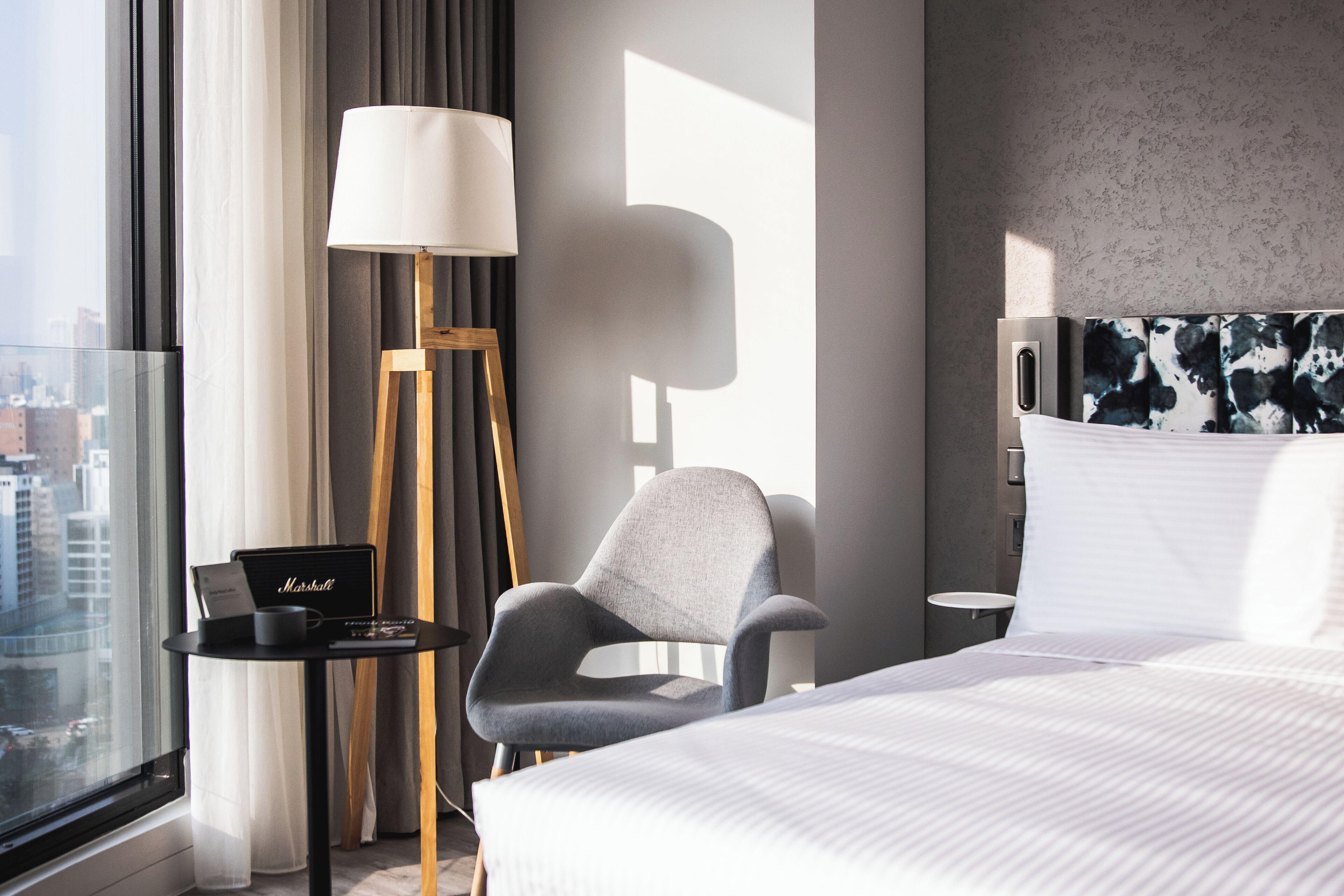 Habitación de hotel con una cama, una silla, una lámpara, una pequeña mesa con una computadora portátil y vistas a la ciudad a través de amplios ventanales.