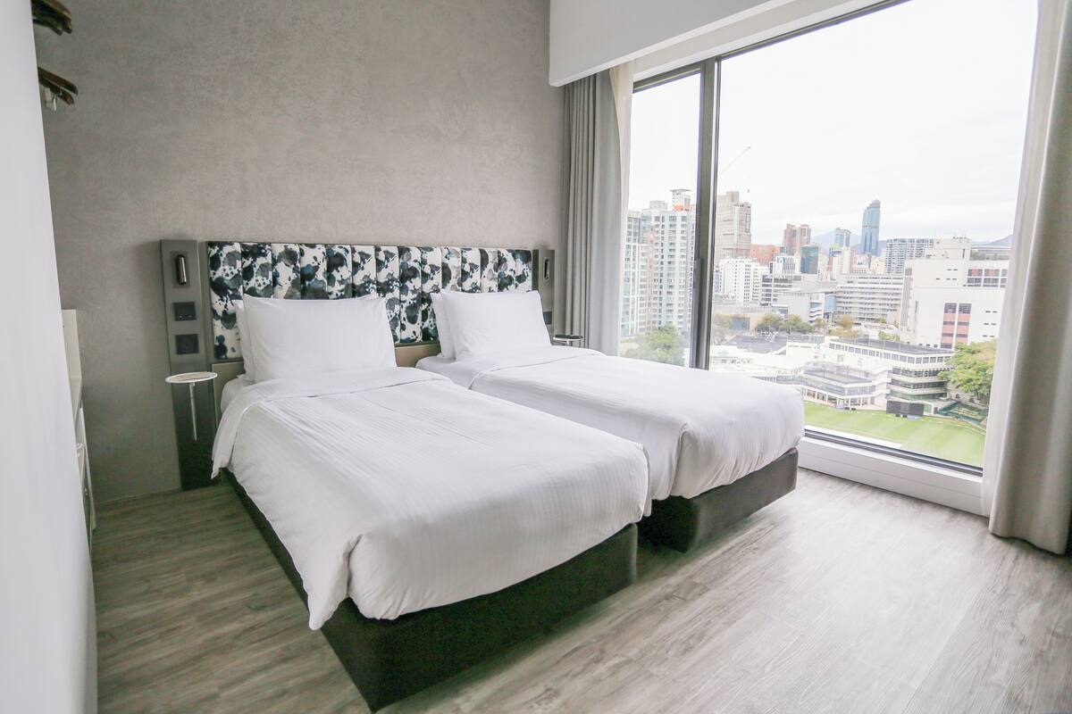 Habitación de hotel con dos camas, un ventanal grande con vistas a la ciudad y un diseño moderno.