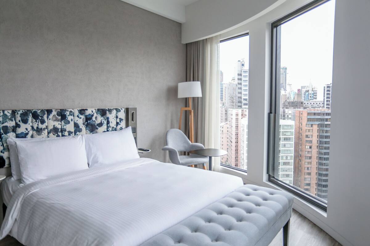 Habitación de hotel moderna con una cama grande, una mesita, una silla y vistas a la ciudad a través de un ventanal.