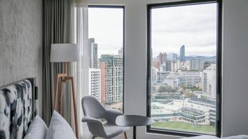 A hotel room with a large window offering a city view, a grey armchair, a bedside table, and a lamp.