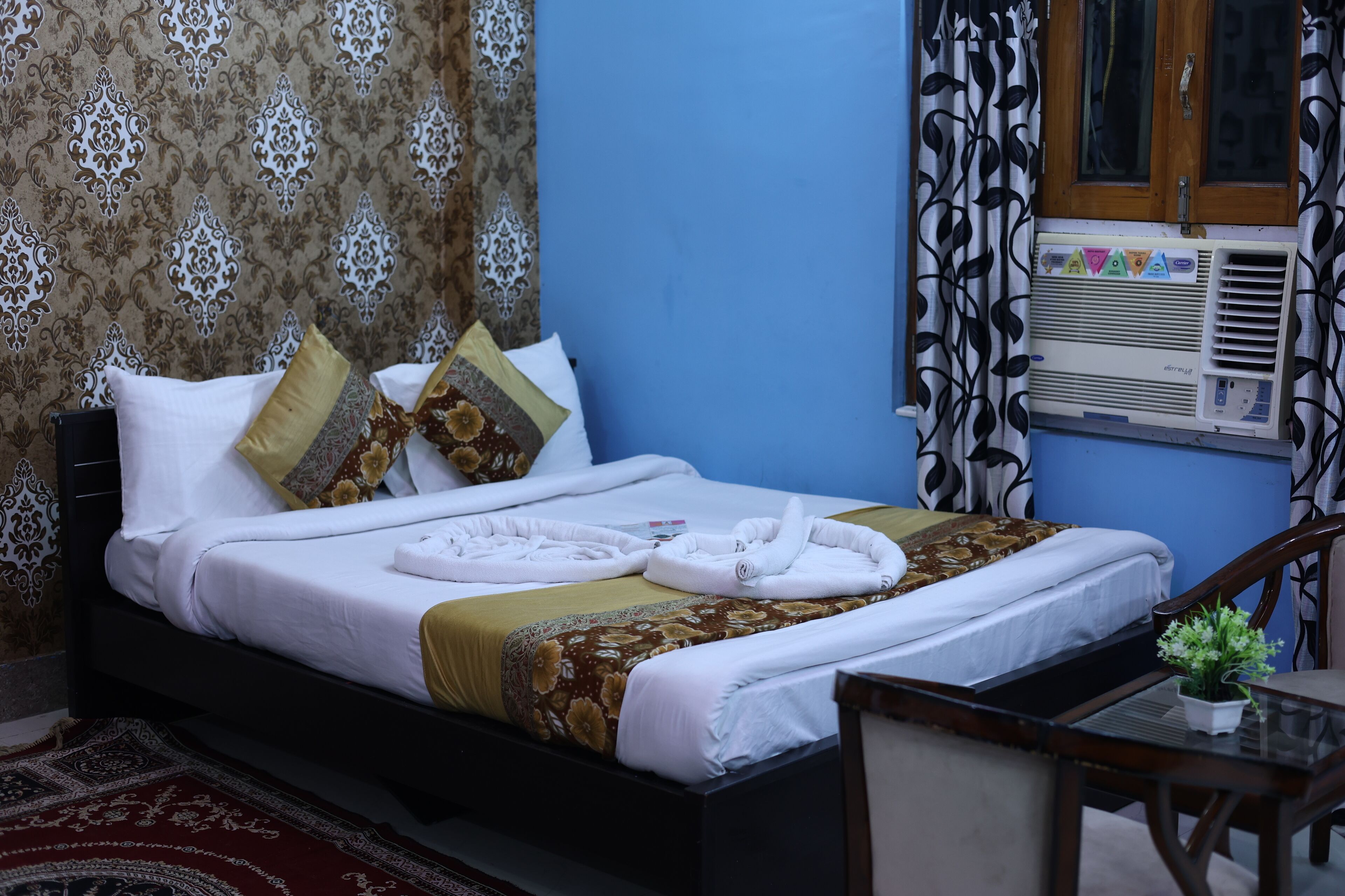 A neatly made bed with white linens and a floral-patterned bedspread, a wooden nightstand, a small table with a potted plant, and a window with a patterned curtain.