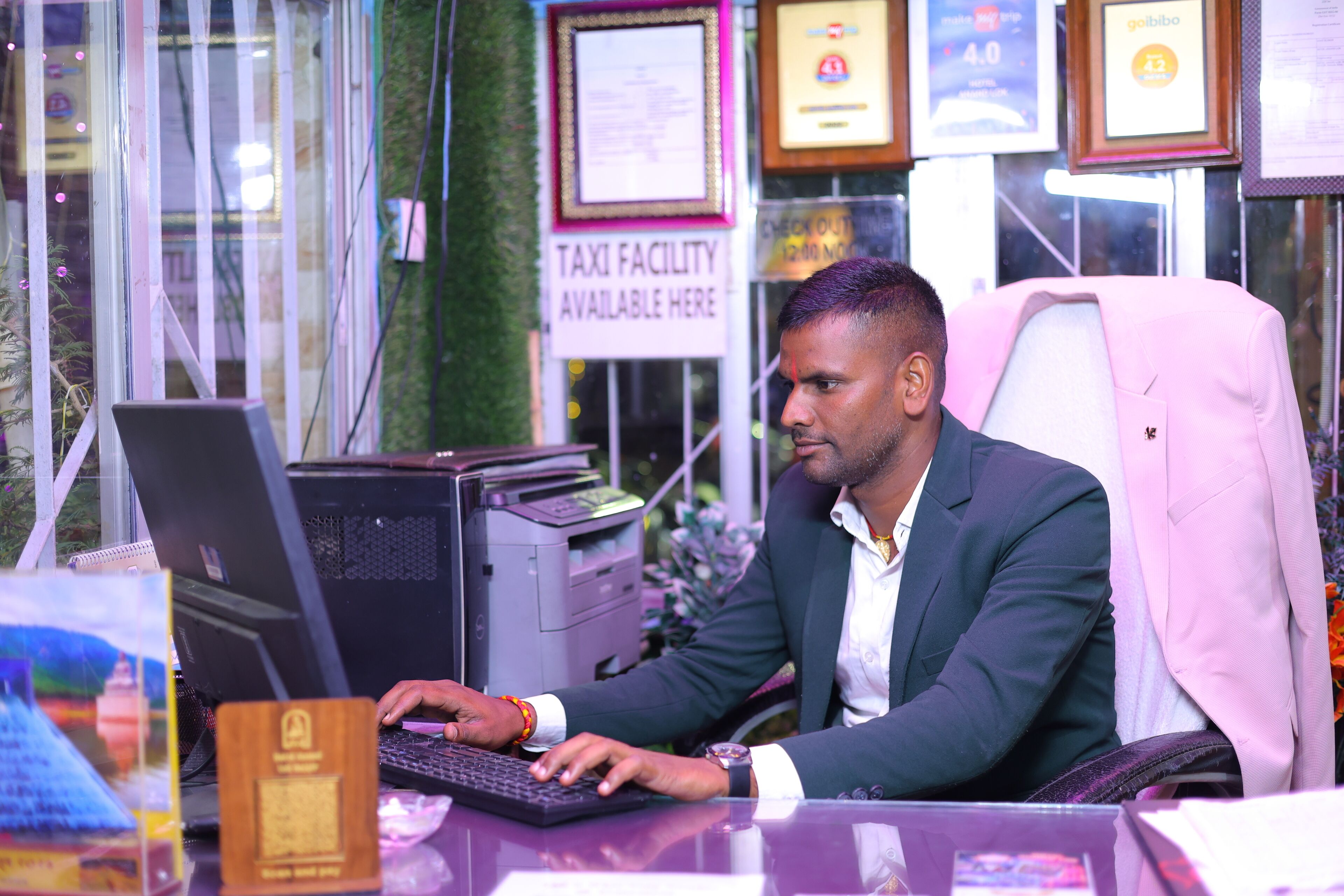 A man working at a desk with a computer, surrounded by framed certificates and a sign indicating a taxi facility.