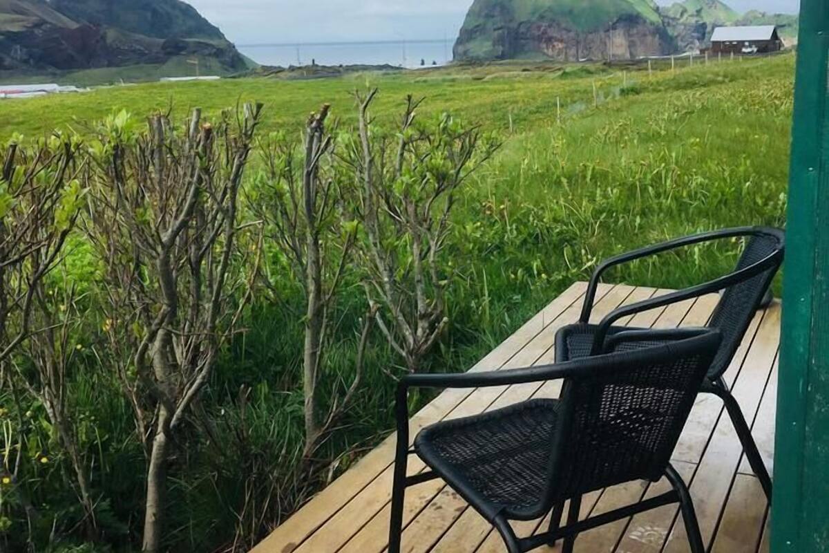 Una terraza de madera con una única silla negra, con vistas a un paisaje cubierto de hierba, colinas y una vista distante del mar.