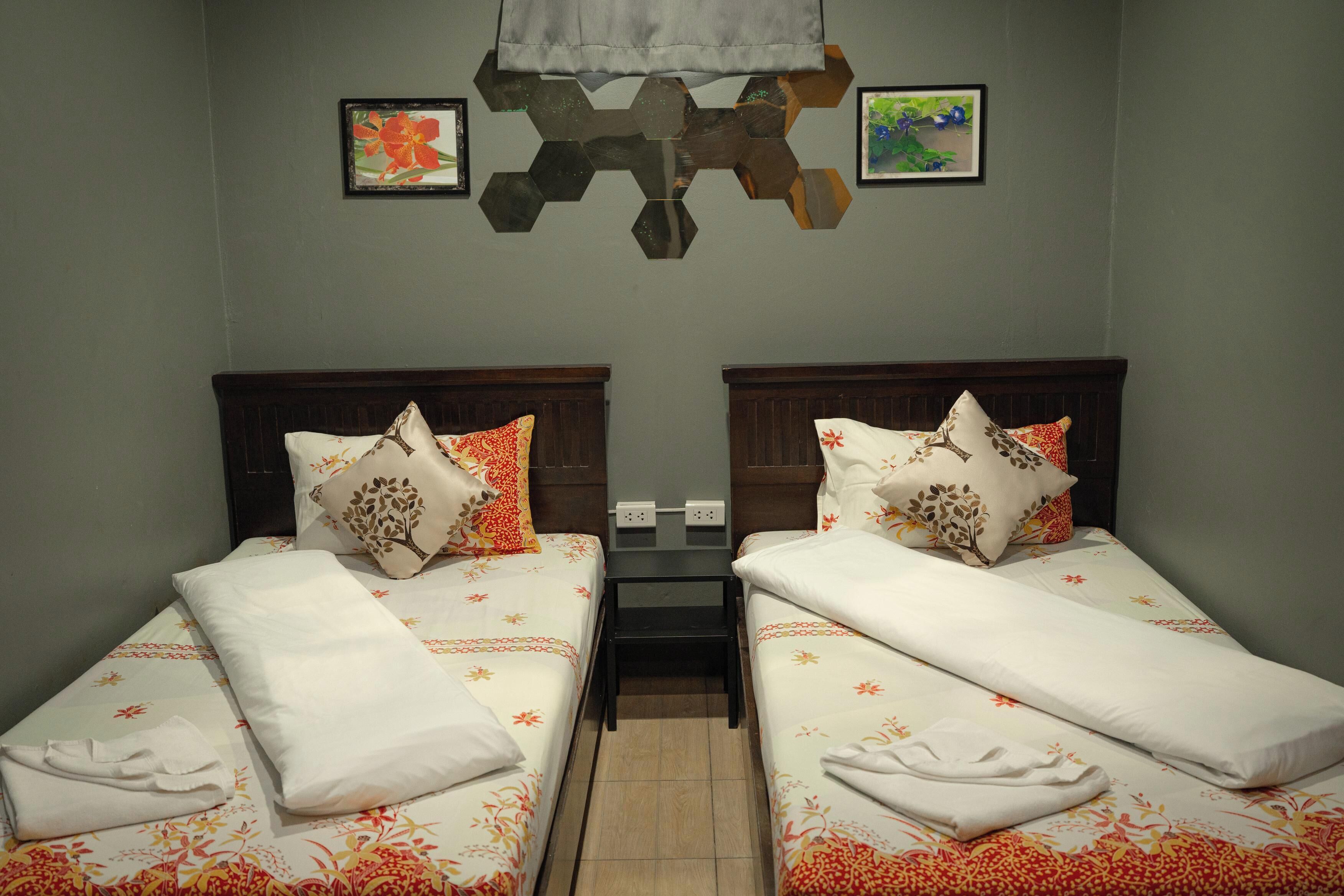 Habitación doble con dos camas, cada una con ropa de cama floral y almohadas a juego. Hay una pequeña mesa entre las camas, y la habitación tiene un diseño sencillo y moderno con una decoración geométrica en la pared sobre las camas.