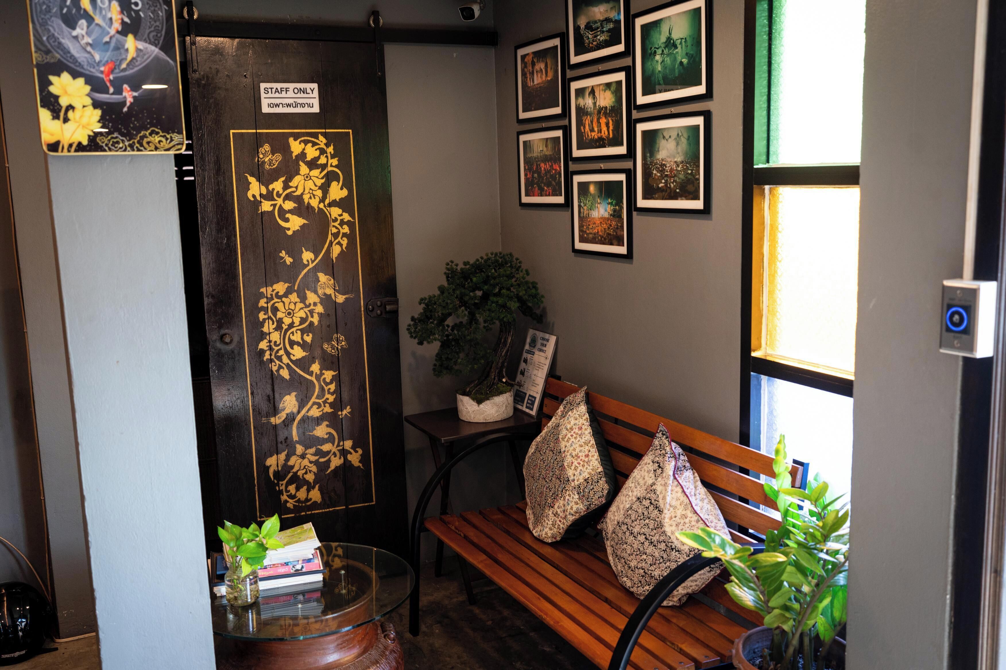 Un área de recepción con una puerta de madera que indica 'STAFF ONLY', un banco con cojines estampados con motivos florales, una pequeña mesa con una planta y cuadros en la pared.