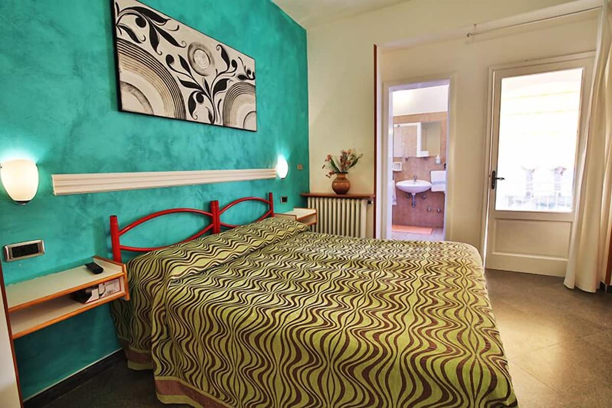 A hotel room with a teal wall, a bed with a patterned bedspread, a red headboard, bedside tables, a nightstand, a wall-mounted artwork, and a view of the bathroom and balcony through an open door.