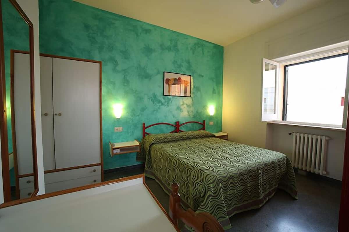 A bedroom with a teal wall, a bed with a patterned bedspread, a wooden wardrobe, bedside lamps, and a window with a view.