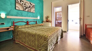 A bedroom with a teal wall, a bed with a patterned bedspread, a red headboard, a bedside table with a lamp, and a door leading to a bathroom. - Albergo 2 Mari (Castrignano del Capo)