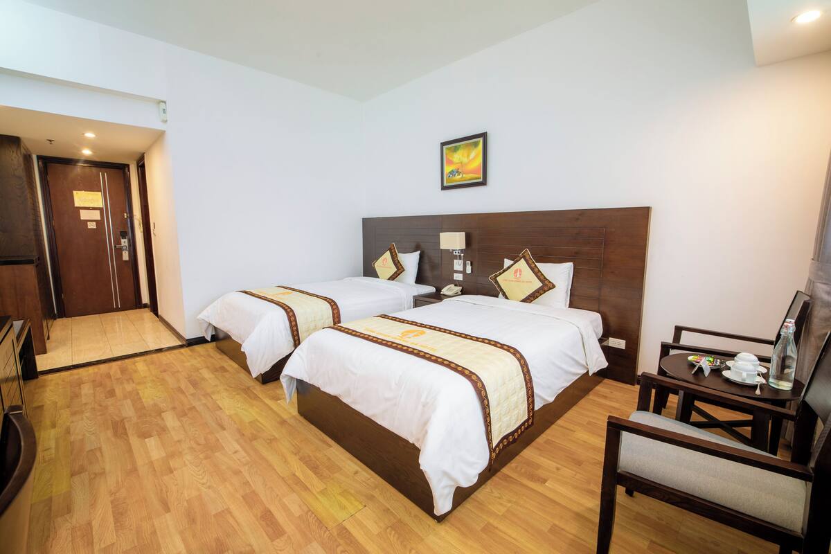 A hotel room with two beds, a desk, a chair, and a small table with a lamp.