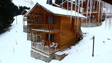 A wooden cabin with a balcony and stone foundation, surrounded by snow and trees.