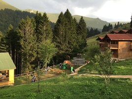 Una cabaña rústica con techo verde, rodeada de un parque infantil y un bosque denso.