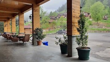 A covered outdoor seating area with wooden pillars and tables, potted plants, and a view of houses in the background.