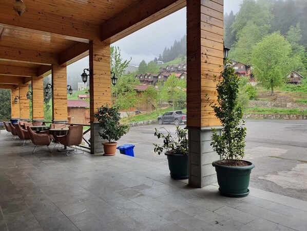 Un espacio de estar al aire libre cubierto, con pilares de madera, mesas, plantas en macetas y vistas a casas al fondo.