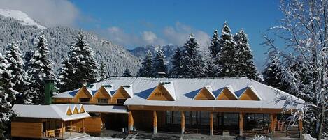 A lodge with a snow-covered roof and wooden exterior, surrounded by snow-covered trees and mountains.