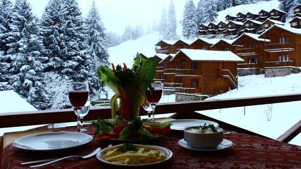 A dining table with a meal set, a glass of red wine, and a view of snow-covered cabins in the background. - Hamsiköy Zitaş Otel & Bungalov & Restoran (Maçka)