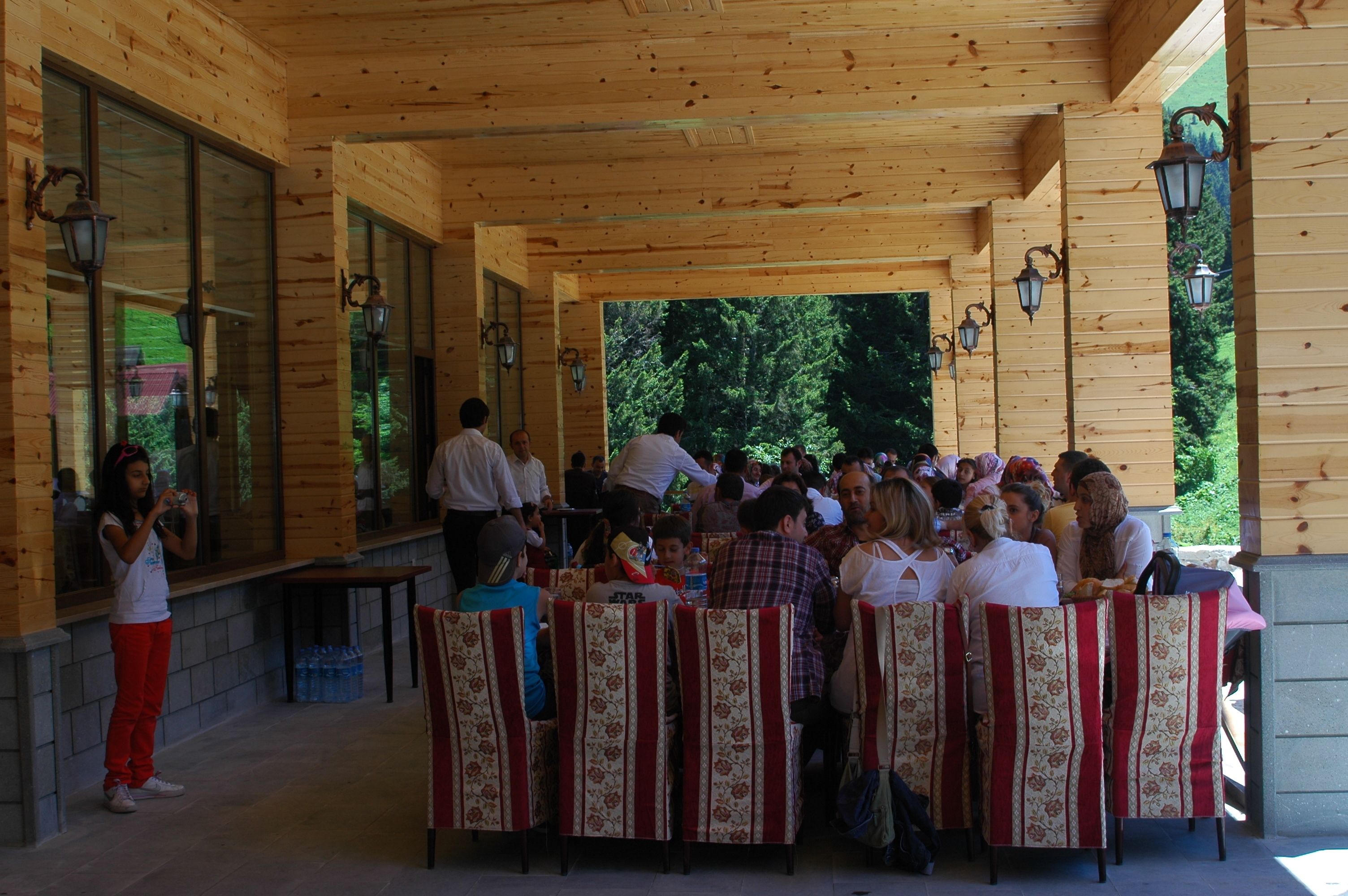 A covered outdoor seating area with wooden walls and large windows, providing a view of the surrounding greenery.