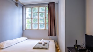 A neatly made bed with a white pillow and a rolled towel, a window with a view of greenery, and a black desk with a television and a device. - K Hotel 14 (Singapore)