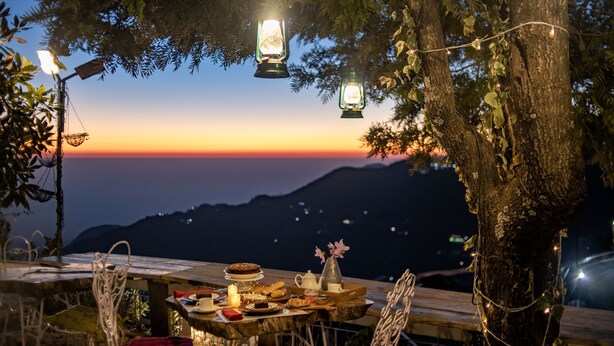 Une table dressée en plein air, avec vue sur le coucher de soleil, des lanternes et des guirlandes lumineuses.