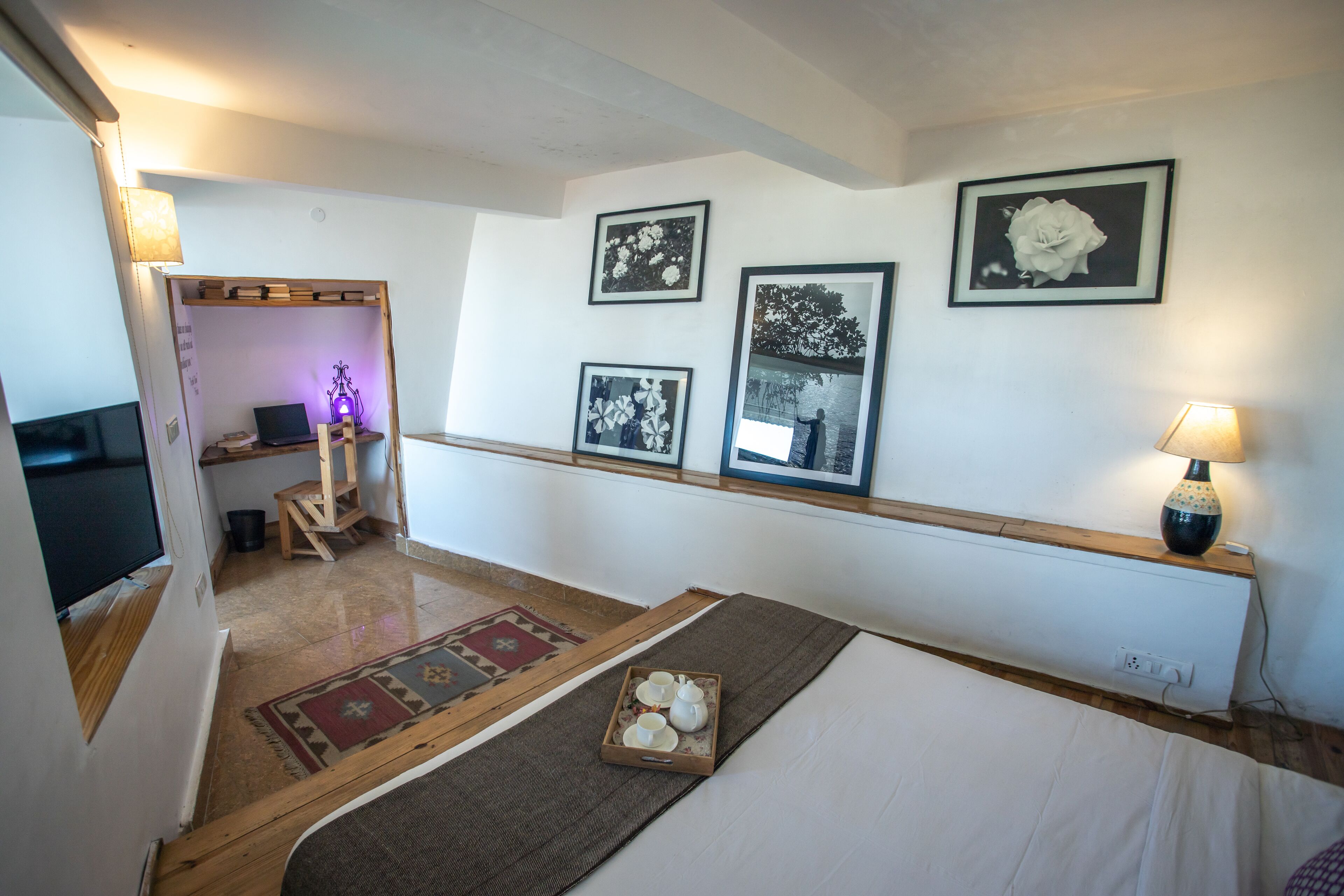 A room with a bed, a desk with a laptop, a small table with teapots, and framed pictures on the wall.