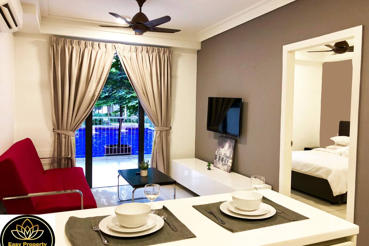 A hotel room with a bed, a TV mounted on the wall, a dining area with a table set for two, and a view of a pool and trees through the window.